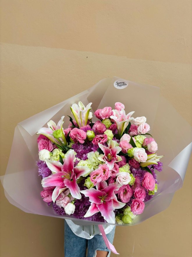 A bouquet of pink stargazer lilies, spray roses, lisianthus, and chrysanthemums wrapped in pastel paper.