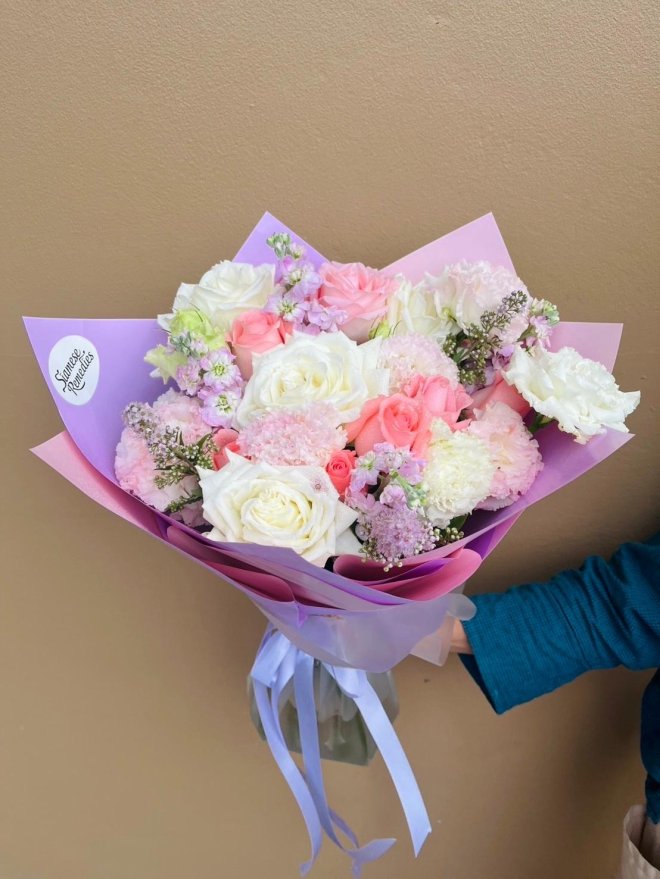 A pastel bouquet with white and pink roses, carnations, lilac stock, and fillers, wrapped in soft lavender and pink paper.