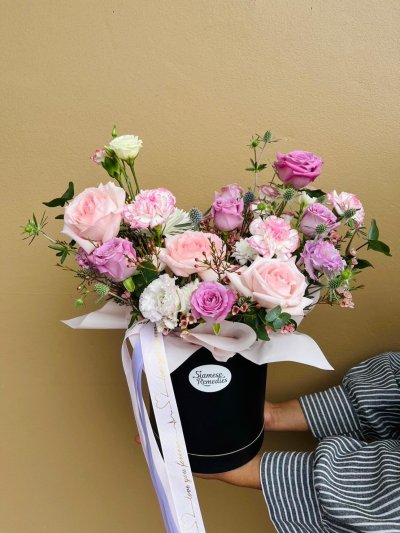 A black hatbox arrangement with pink and lilac roses, carnations, lisianthuses, and wax flowers.