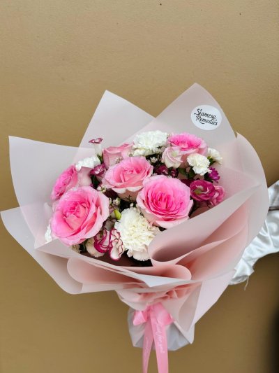 A delicate bouquet with pink roses, white carnations, lisianthus, and wax flowers wrapped in blush pink paper.