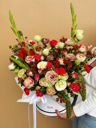 A white hat box filled with red, cream, and purple roses, lisianthuses, carnations, berries, and greenery, tied with white ribbon.