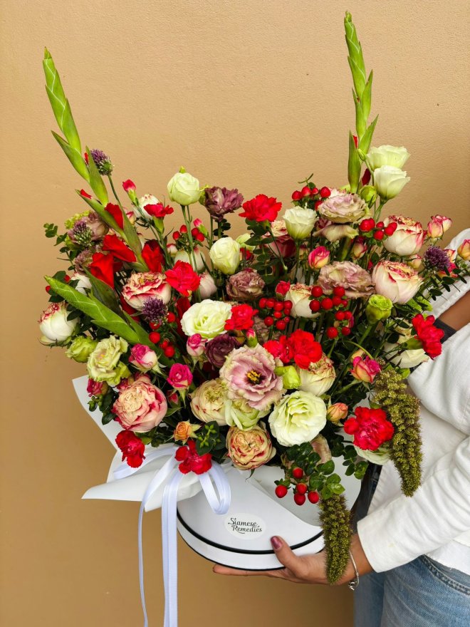 A white hat box filled with red, cream, and purple roses, lisianthuses, carnations, berries, and greenery, tied with white ribbon.