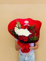Person holding Velvet Noel holiday bouquet with red and white roses, baby's breath, and evergreen sprigs wrapped in red paper with ribbon