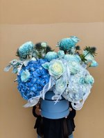 Person holding a large blue hatbox arrangement filled with blue and white hydrangeas, light blue roses, and white ribbons against a beige backgroun...