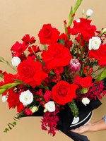 A black hat box arrangement with red roses, white lisianthuses, orchids, protea, and green accents.