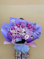 Sutinee bouquet featuring 25 pink, lavender and deep pink roses with blue and purple statice flowers, wrapped in purple paper with ribbons, held ag...