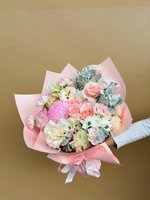 A pastel bouquet of pink roses, carnations, and white flowers wrapped in soft pink paper.