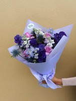 Hand holding a purple-wrapped bouquet featuring deep purple lisianthus, pink roses, white chrysanthemums with textural greenery against a beige bac...