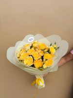 A beautiful hand-tied bouquet of golden yellow ranunculus flowers wrapped in cream paper with yellow ribbon, held against a beige background