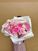 Pink Allure bouquet featuring pink and white lisianthus flowers with baby's breath, wrapped in white paper with gold lettering, held against a beig...