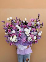 A large arrangement of purple and white roses, lisianthus, and pastel blooms in a white vase with lilac ribbon.