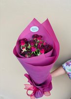 Hand holding Betty bouquet with twelve deep burgundy roses wrapped in purple paper with matching ribbon against neutral background