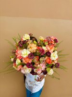 White round box arrangement with orange roses, plum blooms, and lisianthus surrounded by green sprigs.