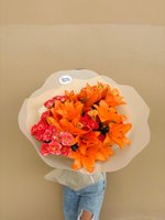 Kezia bouquet featuring bright orange lilies and pink-red roses wrapped in white paper, held against a neutral background