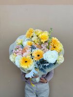Person holding Chesser bouquet with bright yellow-orange gerberas, white and pale yellow lisianthus, pink chrysanthemums, and dusty miller foliage...