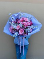 Hand-tied bouquet of pink and purple roses with white stock flowers wrapped in purple paper, held against neutral background