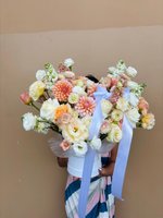 Hand-tied bouquet featuring coral orange dahlias, white and pink ranunculus, white lisianthus, and stock flowers arranged in a white container with...