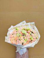 A large bouquet featuring peach, cream, and green roses wrapped in white paper with gold text.