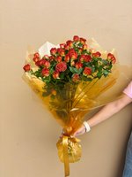 Hand holding a bouquet of vibrant orange-red spray roses wrapped in golden cellophane with yellow ribbon against a neutral background