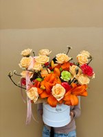 Luxury white box arrangement with vibrant orange lilies, soft peach roses, and burgundy carnations, held by person against neutral background with...