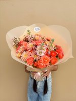 Person holding Regina bouquet with coral-orange ranunculus, roses, pink dahlias and wax flowers wrapped in cream paper against beige background