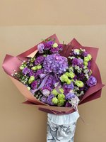 Hand-tied bouquet with purple and lavender hydrangeas, green carnations, and white baby's breath wrapped in burgundy paper, held against a neutral...